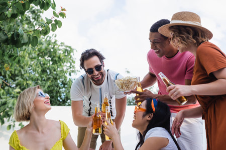 happy multicultural friends clinking bottles of beer near african american man with bowl of chipsの写真素材