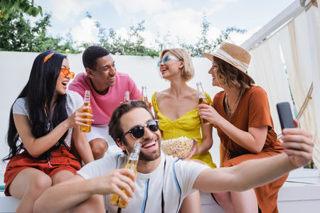 cheerful man in sunglasses taking selfie near interracial friends drinking beerの写真素材