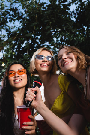 Low angle view of smiling multiethnic women with plastic cup holding microphone outdoors during partyの写真素材