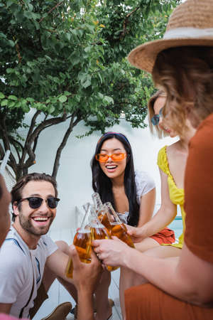 excited multiethnic friends clinking with bottles of beer near green tree in patioの写真素材