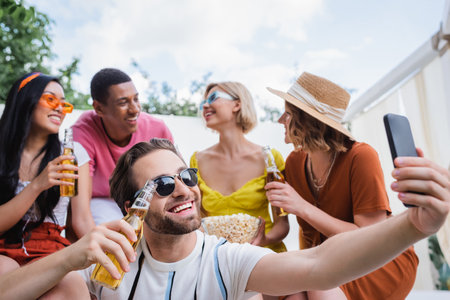 joyful man taking selfie on smartphone during summer party with blurred multiethnic friendsの写真素材