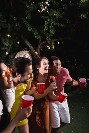 Smiling interracial young people singing karaoke and holding plastic cups outdoorsの写真素材
