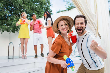excited man showing triumph gesture near woman with water pistol during summer party with blurred...