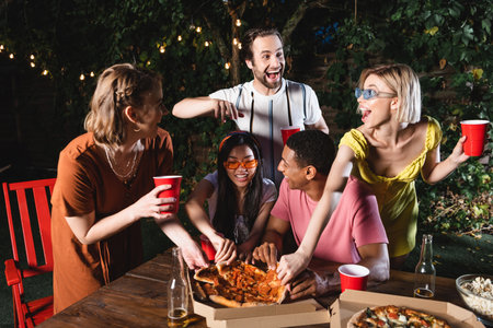 Young multiethnic friends taking pizza near bottles of beer outdoors
