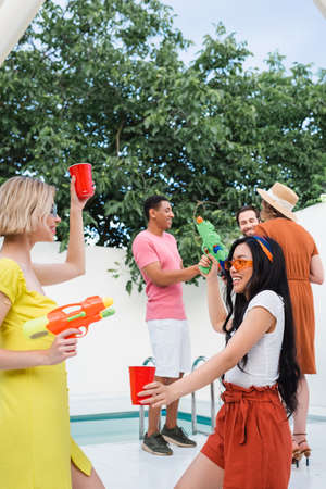 interracial women playing with water pistols near multiethnic friends on blurred background