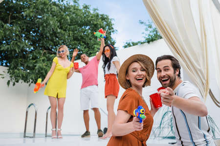 laughing man pointing at camera near woman in straw hat and blurred multiethnic friends with water...