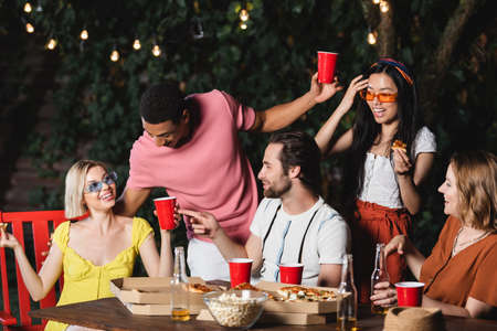 Young man pointing with finger at friend with plastic cup near multiethnic people and food outdoorsの写真素材