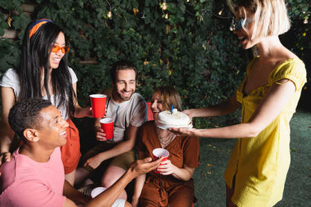 Woman holding birthday cake near african american friend and people with plastic cups outdoors