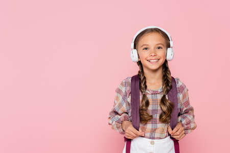 Smiling schoolgirl in headphones holding backpack isolated on pink