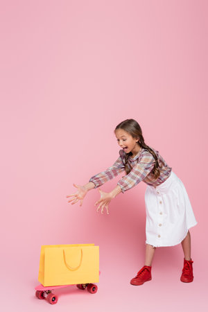 excited girl with open mouth and outstretched hands near yellow shopping bag and penny board on pink backgroundの写真素材