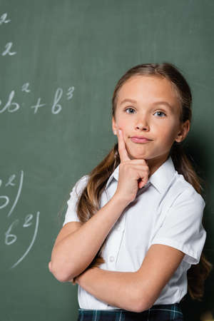 thoughtful schoolkid looking at camera near chalkboard in school