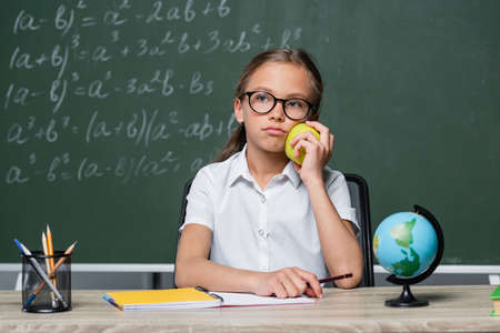displeased schoolgirl holding apple near globe and notebook on deskの写真素材