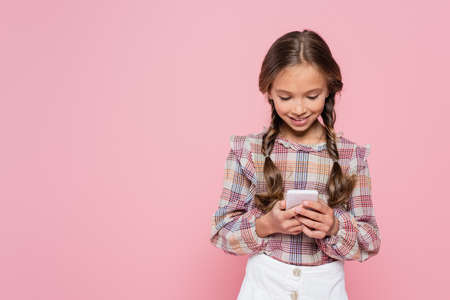 smiling girl in checkered blouse messaging on smartphone isolated on pinkの写真素材
