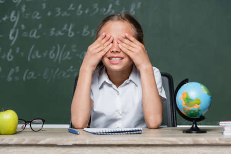 cheerful schoolgirl covering eyes with hands near globe and notebook on desk