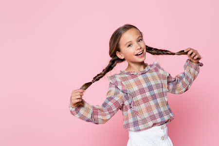 cheerful girl in plaid blouse touching pigtails while looking at camera isolated on pink