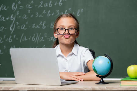 playful schoolgirl with pencil between lips and nose sitting near laptop, globe and blurred chalkboardの写真素材