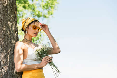 trendy woman in sunglasses and yellow headscarf holding flowers and standing near tree trunkの写真素材