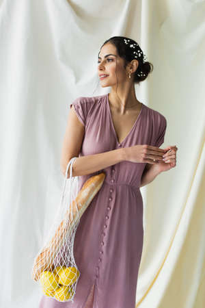 happy woman holding reusable string bag with lemons and baguette on white