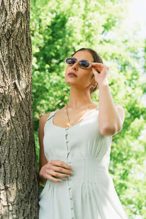 brunette woman in dress adjusting sunglasses near tree trunk