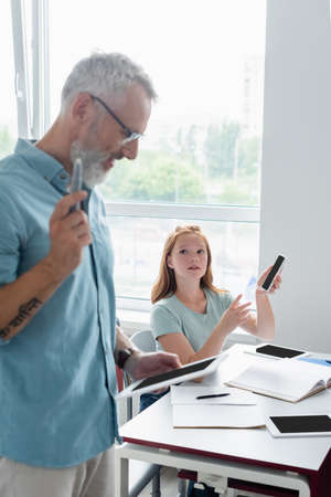 Schoolgirl holding smartphone near blurred teacher with digital tabletの写真素材