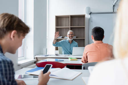 Cheerful teacher waving hand at pupils on blurred foreground in classroom