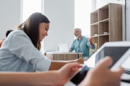 Smiling teacher using laptop and pointing with finger near blurred pupils