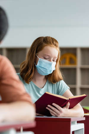 Schoolgirl in medical mask reading book in classroomの写真素材