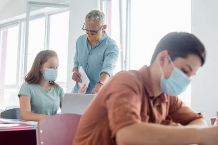 Mature teacher in medical mask standing near schoolgirl pointing at laptop during lesson