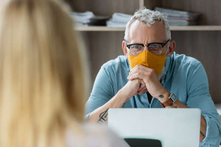 Middle aged teacher in protective mask looking at blurred pupil near laptop in classroom