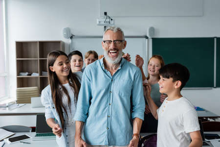 cheerful multiethnic classmates near happy teacher laughing with closed eyes in classroomの写真素材