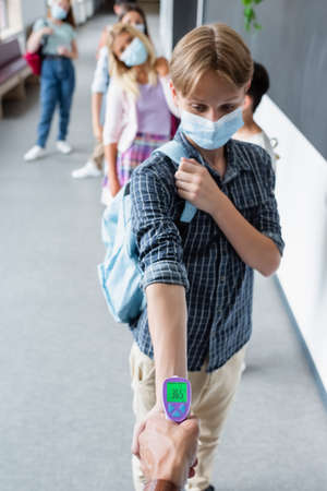 teenage boy in medical mask near teacher measuring his temperature with pyrometerの写真素材