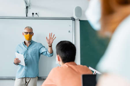 Teacher in protective mask holding digital tablet and showing palm of hand near pupils