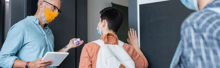teacher with pyrometer and digital tablet inviting schoolboy in medical mask into classroom, banner