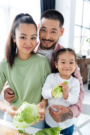 happy asian man and woman looking at camera and smiling near daughter with fresh lettuce