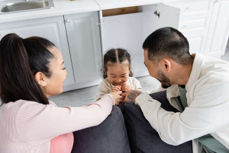 happy asian kid hiding in kitchen cabinet near blurred parents on sofa