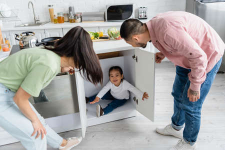 happy toddler asian kid hiding in kitchen cabinet near parentsの写真素材