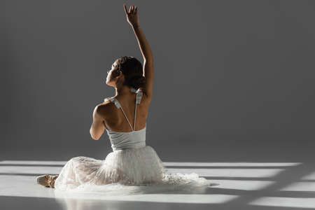 Ballerina raising hand while sitting in sunlight on gray background with sunlightの写真素材