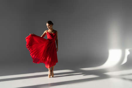 Ballerina in pointe shoes holding red dress on gray background with sunlightの写真素材