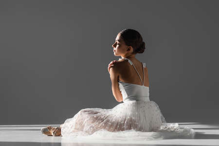 Side view of young ballerina touching shoulder on gray background with sunlight