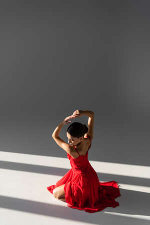 Pretty dancer in red dress posing on gray background with sunlightの写真素材