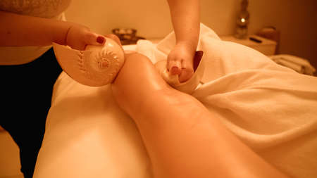 cropped view of therapist massaging leg of young client with sea shells in wellness centerの写真素材