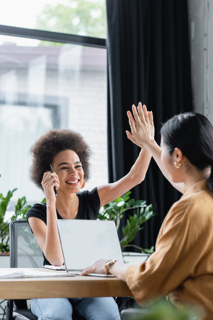 happy interracial businesswomen giving high five while working near laptop at workplace
