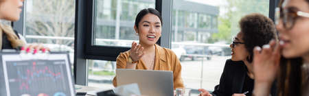 smiling asian woman pointing with hand while talking to blurred multiethnic colleagues, banner