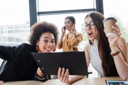 excited interracial managers looking at clipboard near while happy asian businesswoman talking on smartphoneの写真素材