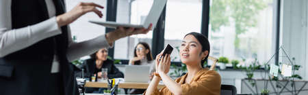 asian businesswoman covering smartphone with hand near colleague using laptop on blurred foreground, bannerの写真素材