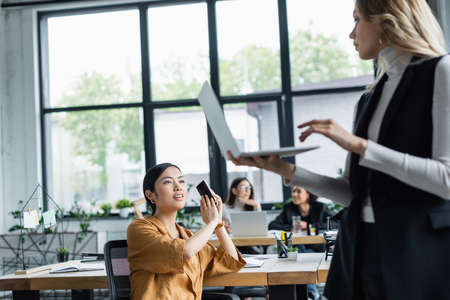 asian businesswoman covering smartphone with hand while talking to blurred colleague with laptopの写真素材