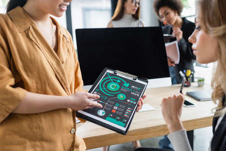manager pointing at clipboard with analytics near blurred colleague in office