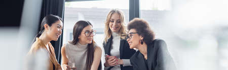 cheerful multicultural businesswomen talking in office on blurred foreground, bannerの写真素材