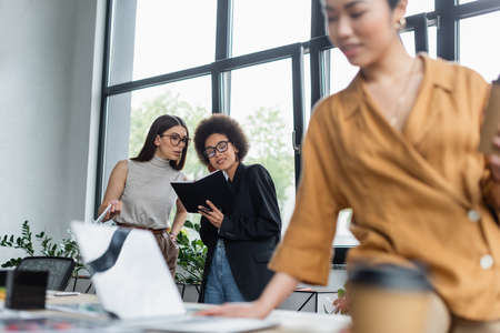 interracial colleagues in eyeglasses looking at notebook near blurred asian businesswomanの写真素材