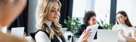 smiling businesswoman looking at digital tablet near blurred multiethnic colleagues in office, bannerの写真素材